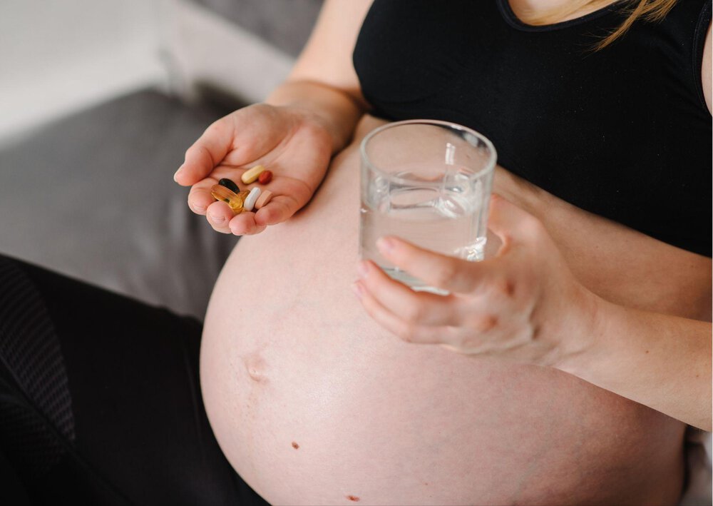 Kobieta w zaawansowanej ciąży trzyma szklankę i kilka tabletek, a w tym witaminę D3